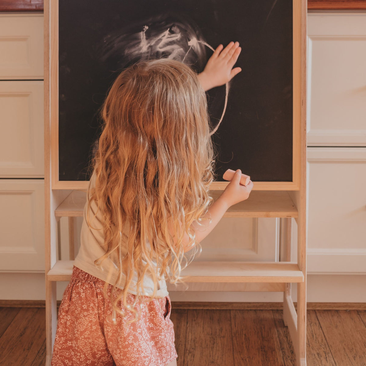 Image of a child with her back to camera, long blonde hair, drawing on with chalk on a handmade wooden chalkboard