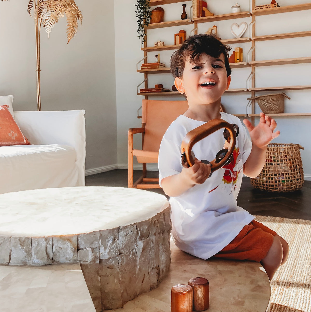 an image of a smiling child with dark hair sitting on a natural table top playing a wooden tambourine instruments for kids