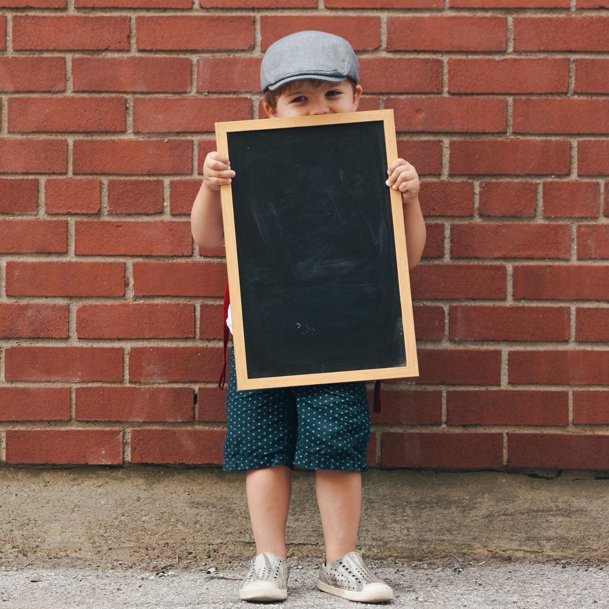 Image of a small child holding a wooden chalkboard standing against a red brick wall and wearing a hat