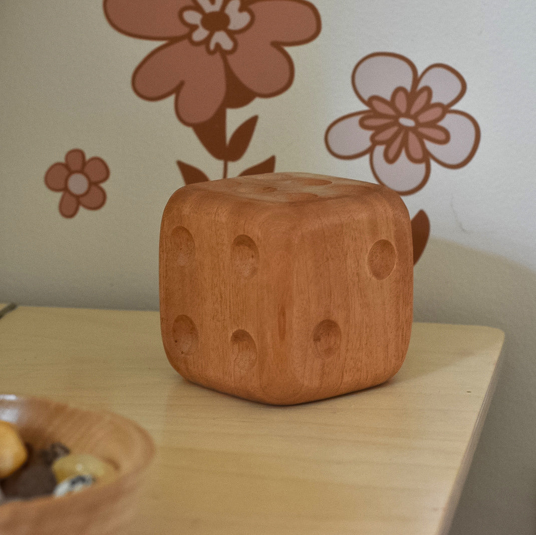 Image of a large wooden dice on a timber table with a wooden bowl and flowers on the wall in the background