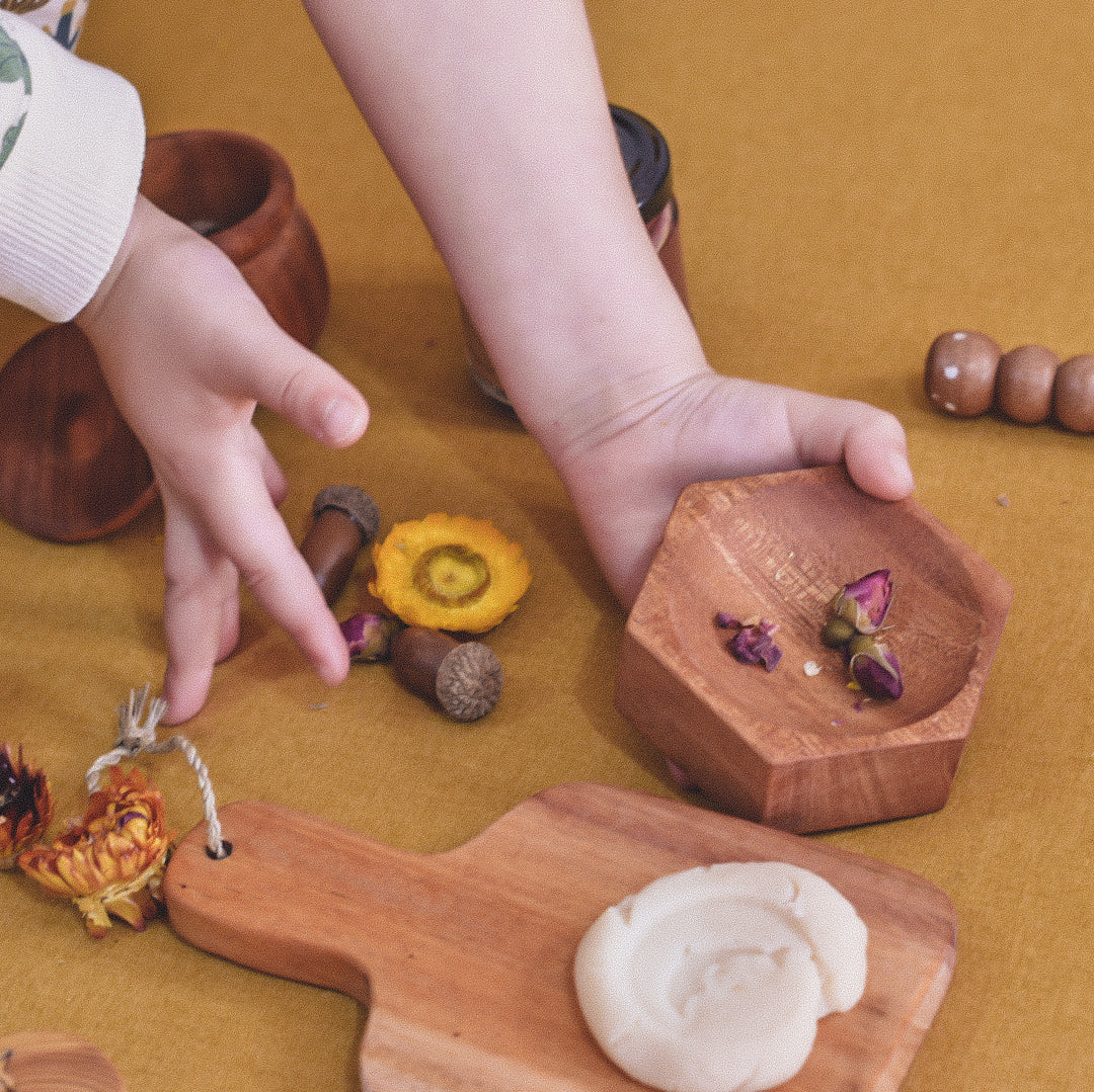 a solid mahogany wooden honeycomb dish filled with dried flowers in child's hand on a yellow background surrounded by a small wooden board with play dough 