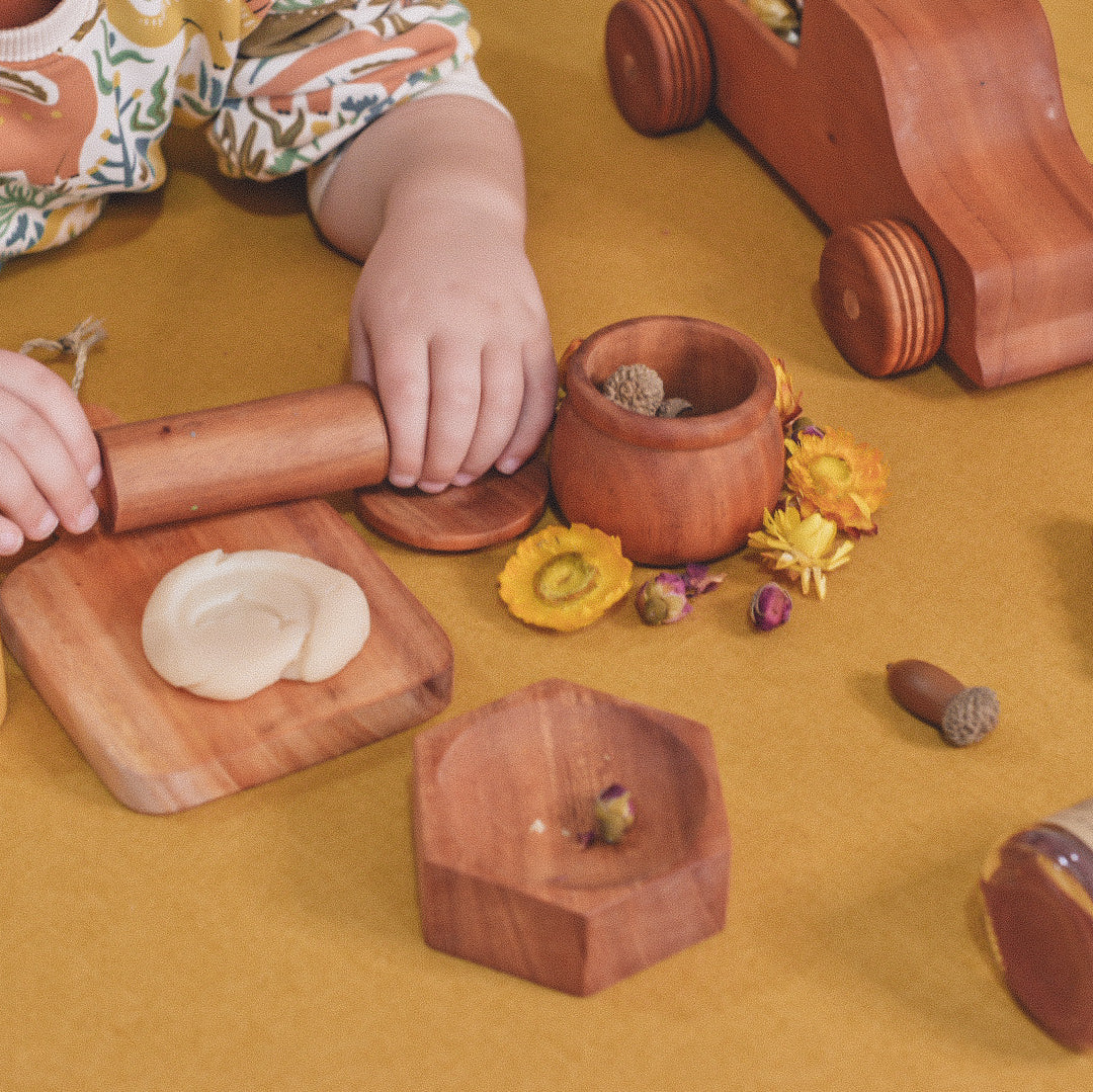 wooden play dough accessories and child's hands on a yellow background with a solid mahogany wooden honeycomb dish filled with dried flowers