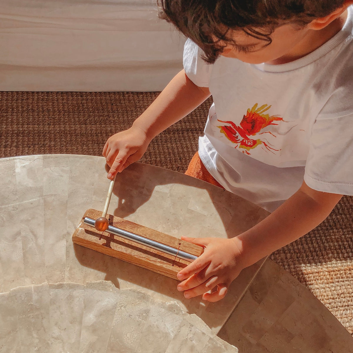 an image of a child wearing a white printed shirt sitting at a light coloured table playing a wooden chime bar instrument and there is a jute rug on the floor in the background