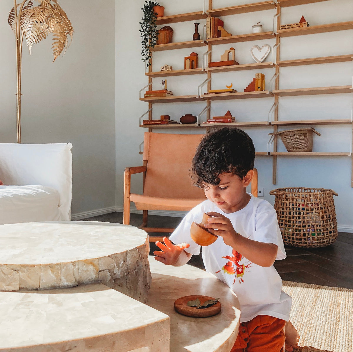 an image of a child wearing a white print shirt and red shorts kneeling at a natural table playing with a wooden mortar and pestle herb crusher they are holding the mushroom shaped handle and they are crushing a leaf