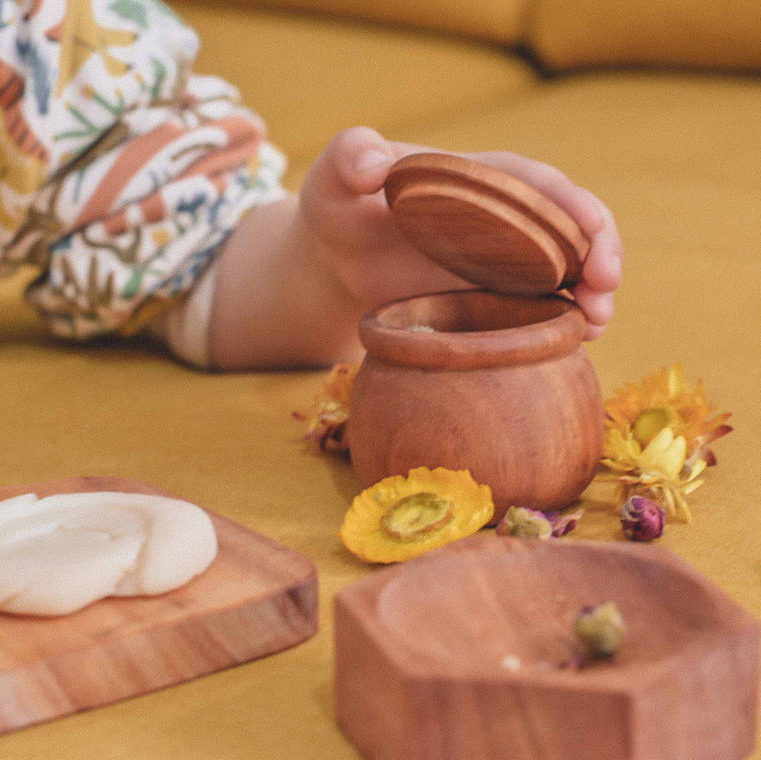 a child's hand lifting the lid of a wooden jar on a yellow background scattered with dried flowers and a solid mahogany wooden honeycomb dish filled with dried flowers