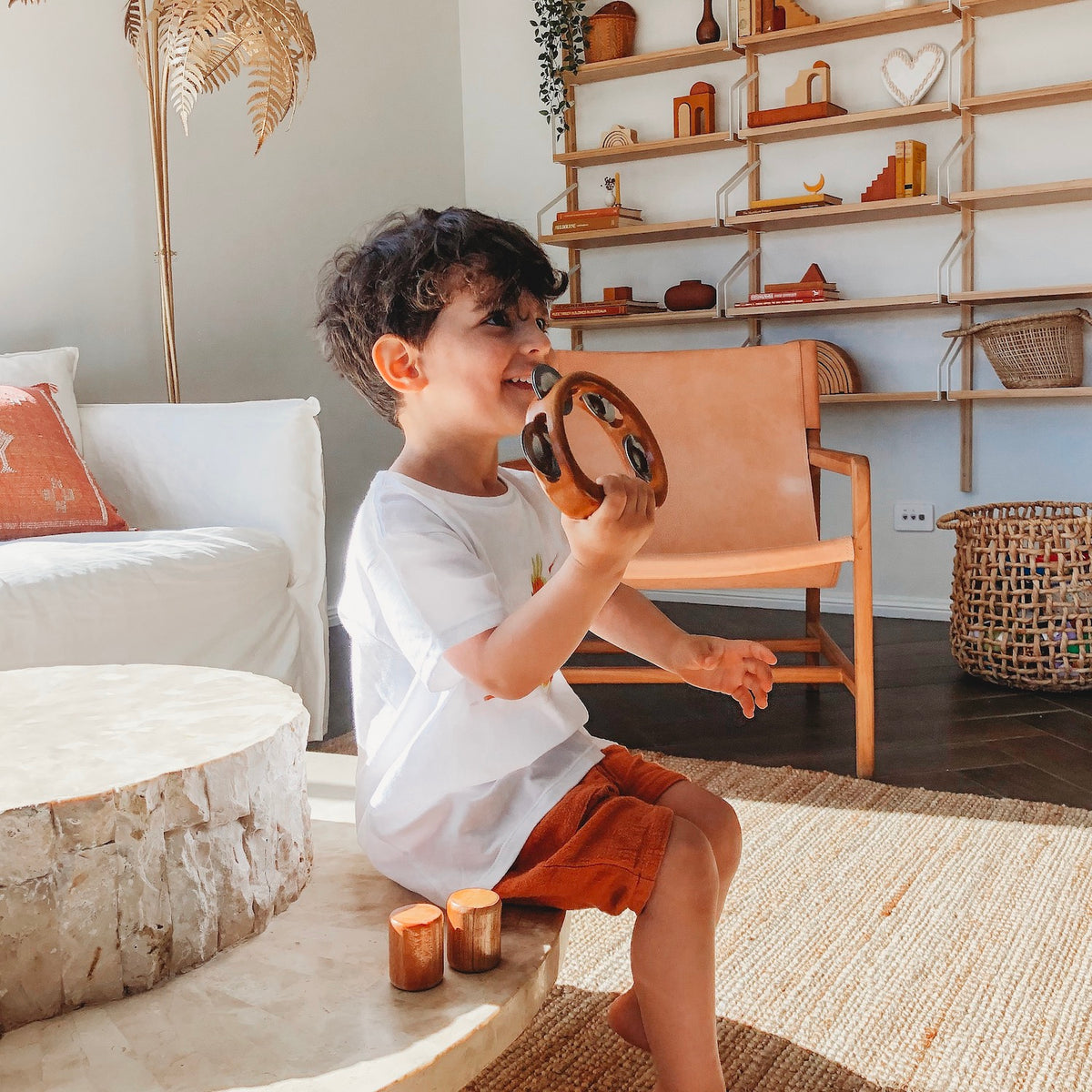 an image of a smiling child with dark hair sitting on a natural table top playing a wooden tambourine instruments for kids