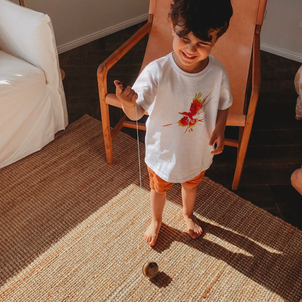 an image of a child with dark hair wearing a white printed tshirt and red shorts standing in front of a  tan coloured chair playing with a wooden yo yo