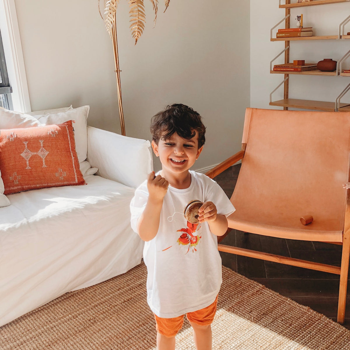 an image of a smiling child with dark hair wearing a white printed tshirt and red shorts standing in front of a  tan coloured chair and white couch playing with a wooden yo yo